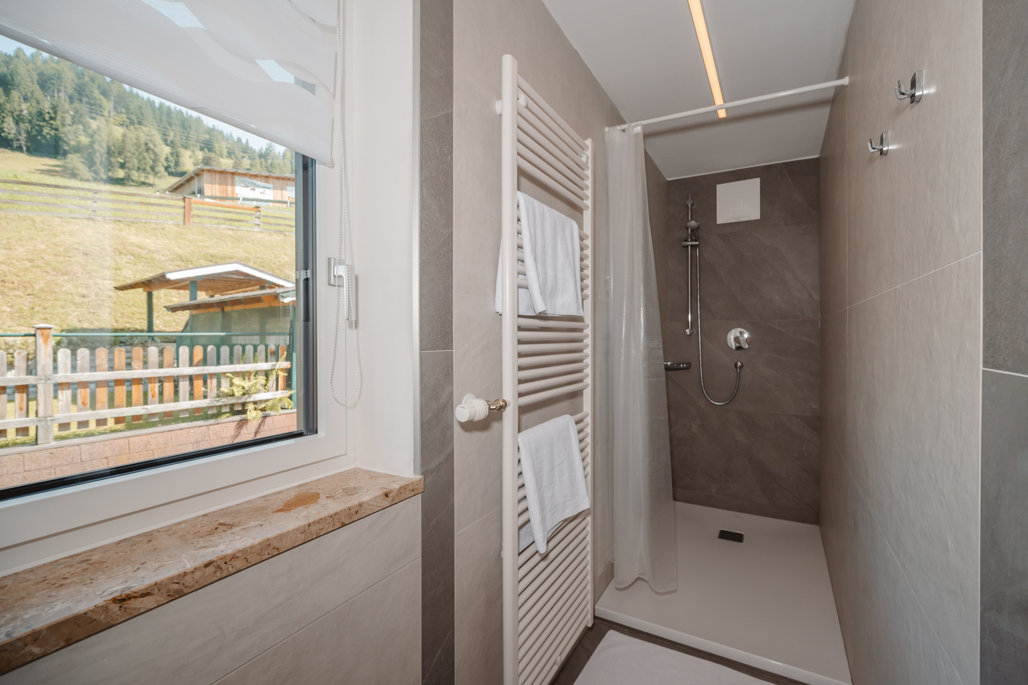 MonteurZimmer: Ein modernes Badezimmer mit einer begehbaren Dusche und einem großen Fenster. Im Ausblick des Fensters sieht man eine grüne Landschaft mit einem Gartenhaus. - Haus Ortsblick